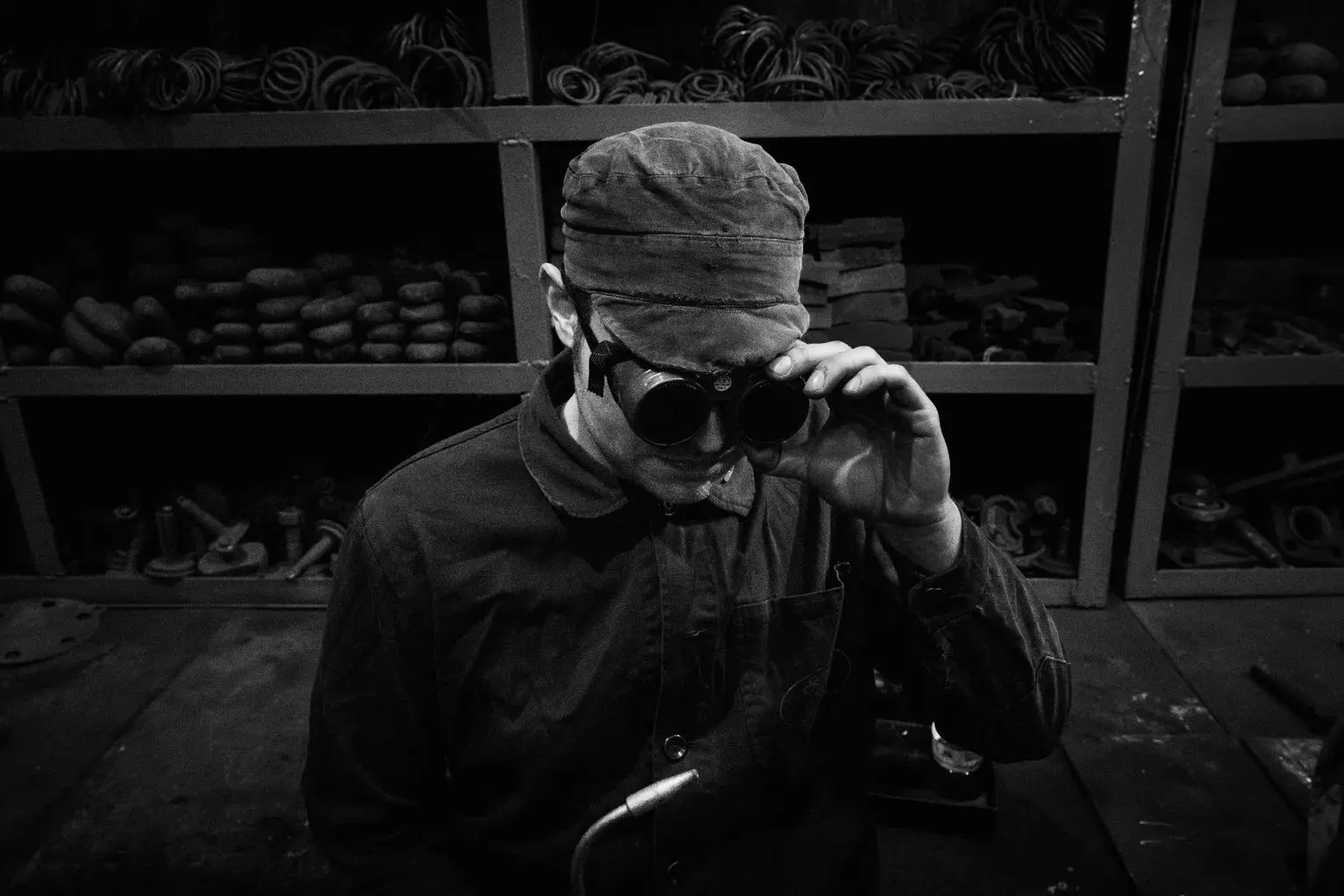 Retrato en blanco y negro de un trabajador ferroviario con gorra y gafas protectoras frente a estanterías de piezas.