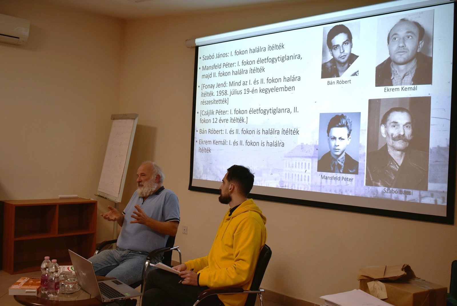 László Eörsi durante una conferencia, junto a una proyección sobre participantes y represaliados de 1956.