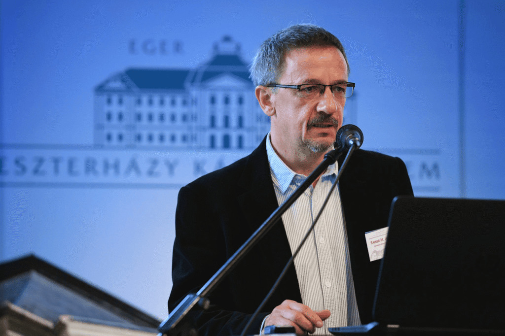 El historiador János M. Rainer habla ante un micrófono, con la imagen de la Universidad Católica Eszterházy Károly al fondo.