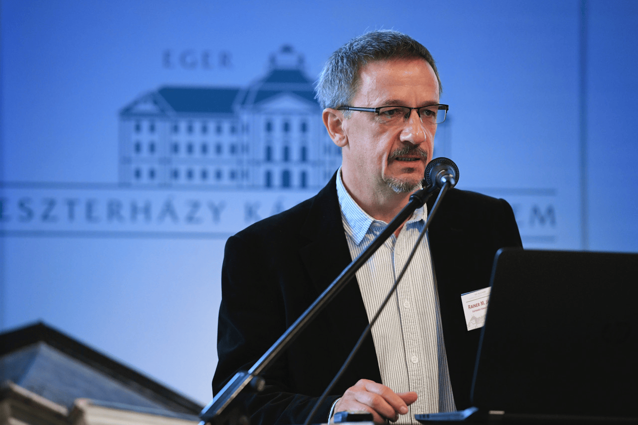 Rainer M. János történész mikrofon előtt beszél; a háttérben az Eszterházy Károly Egyetem épületének képe látható.